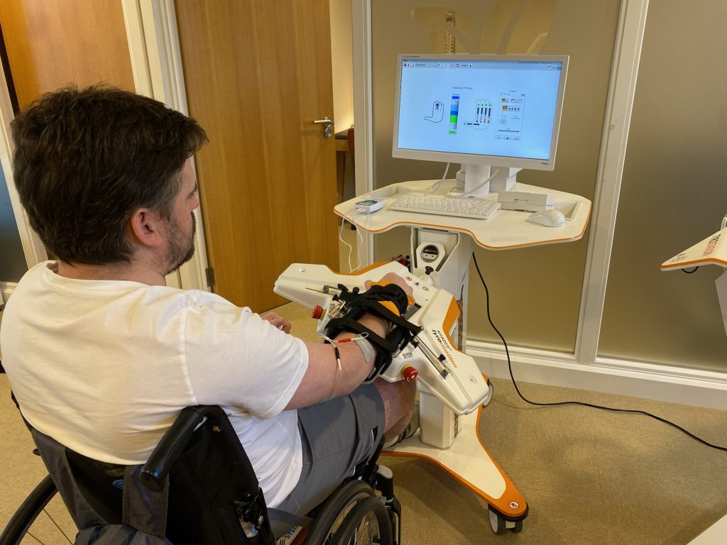 sid is sat using upper limb rehab device the amadeo on his right hand