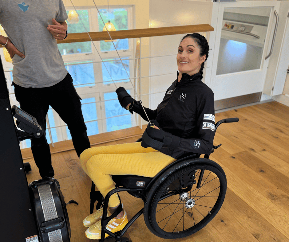 marianne sits in her wheelchair whilst using the skiErg. she is wearing a black sports top with RS logo on it, yellow leggings and active hands gloves.