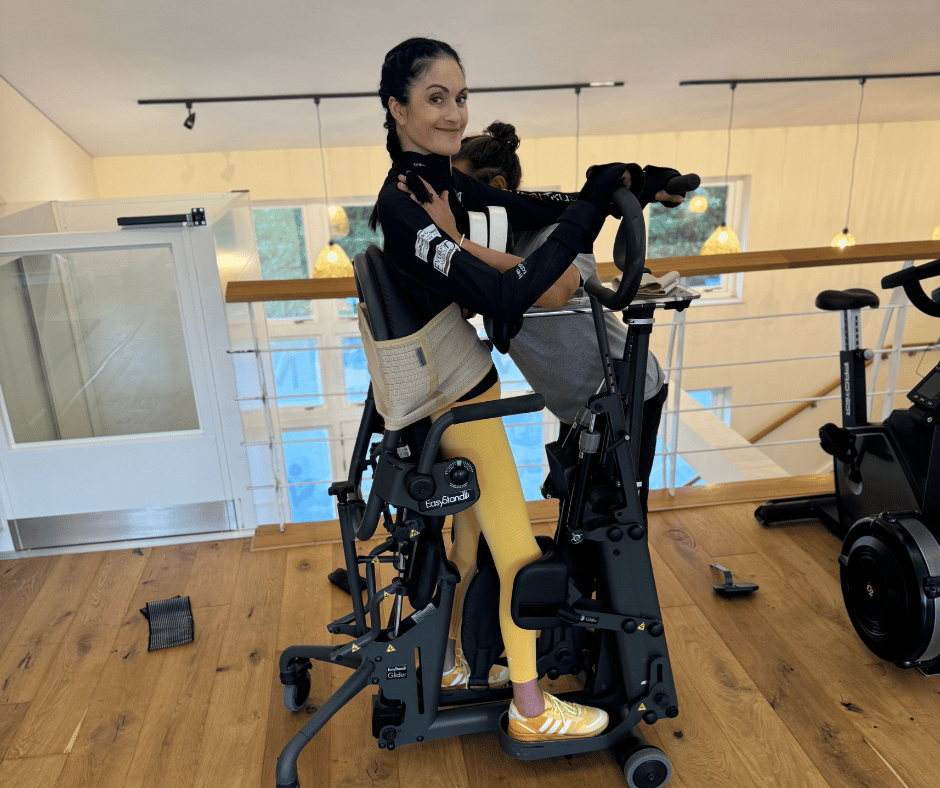 Marianne is stood in the EasyStand Glider. She is smiling at the camera and wearing a blck long sleeved top and yellow leggings.