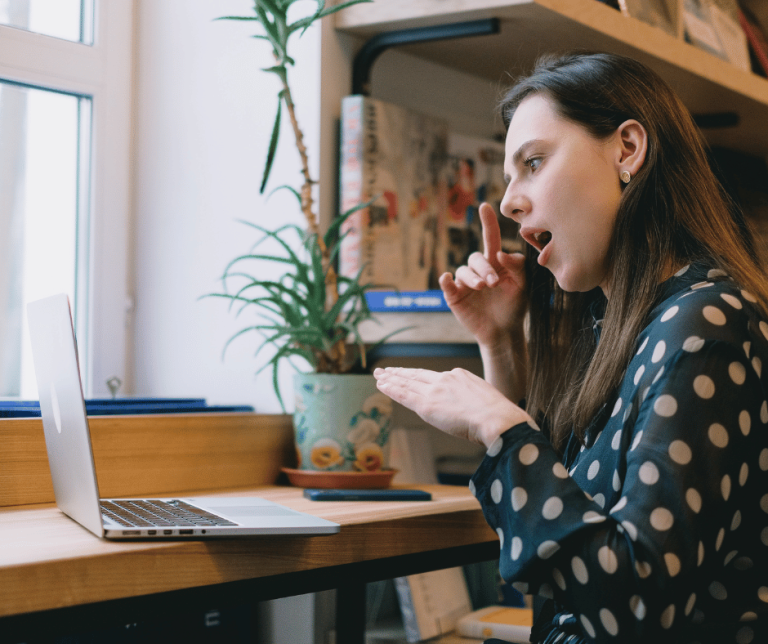 woman sat in front of her laptop signing with her hands and her mouth open. she is wearing a polka dot top